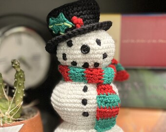 May include: A handmade crochet snowman with a black top hat adorned with holly and berries. The snowman has a red and green striped scarf and black button eyes. A festive winter decoration.