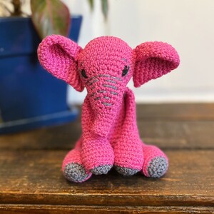 Könnte beinhalten: Ein handgehäkelter, rosa Elefant mit grauen Akzenten. Der Elefant hat große Ohren, einen Rüssel und graue Nahtdetails. Die Füße sind grau. Der Elefant sitzt auf einer Holzoberfläche.