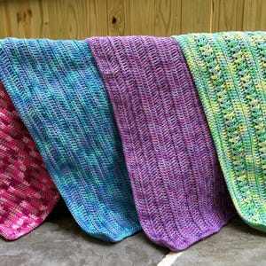 May include: Four colorful, handmade crochet blankets are displayed. The blankets are in shades of pink, blue, purple, and green. Each blanket has a unique stitch pattern, with a textured appearance. The blankets are folded over a wooden surface.