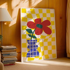 May include: A framed art print featuring a large red flower with green leaves in a blue vase against a yellow and white checkered background. The artwork is displayed in a room with a lamp, books, and a chair.