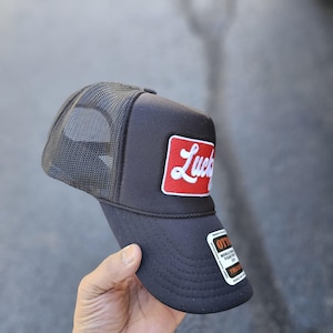 May include: A black trucker hat featuring a mesh back and a rectangular red patch on the front panel. The patch displays the word Lucky in white cursive script. A black and white sticker is attached to the curved brim with visible text.