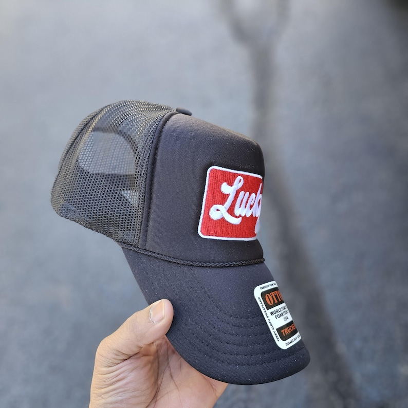 May include: A black trucker hat featuring a mesh back and a rectangular red patch on the front panel. The patch displays the word Lucky in white cursive script. A black and white sticker is attached to the curved brim with visible text.