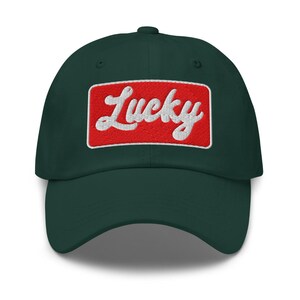 Może przedstawiać: Ciemnozielona czapka bejsbolowa z zakrzywionym daszkiem. Na czapce z przodu znajduje się napis "Lucky" wyszyty białą czcionką w czerwonym prostokątnym polu.