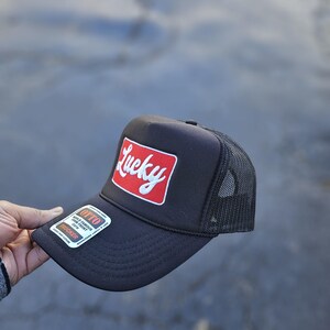 May include: A black trucker hat featuring a rectangular red patch on the front with the word Lucky in white script. The hat has a mesh back, a foam front panel, and a sticker on the brim that reads Otto World Famous Foam Front OSFM Trucker.