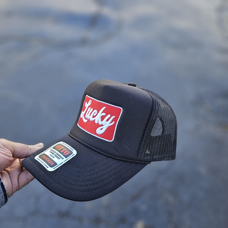 May include: A black trucker hat featuring a rectangular red patch on the front with the word Lucky in white script. The hat has a mesh back, a foam front panel, and a sticker on the brim that reads Otto World Famous Foam Front OSFM Trucker.