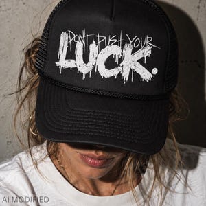 May include: A black trucker hat featuring a mesh back and a solid front panel. The front displays the white text "DON'T PUSH YOUR LUCK." in a distressed, dripping font style. The cap includes a black brim with horizontal stitching and a cord detail.