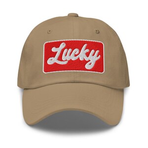 Może przedstawiać: Beżowa czapka z daszkiem z zakrzywionym daszkiem. Czapka posiada haftowany czerwony prostokąt z napisem "Lucky" w białym skrypcie. Czapka ma sześciopanelową konstrukcję z guzikiem na g&oacute;rze.