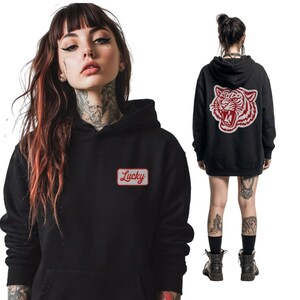 May include: Black hoodie with a red and white tiger graphic on the back and a "Lucky" patch on the front. The hoodie has a drawstring hood and a kangaroo pocket. The model is wearing black boots.