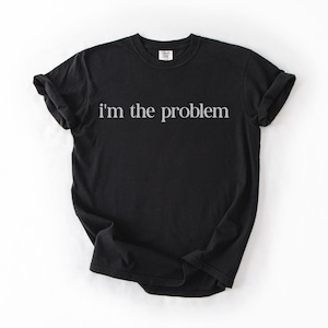May include: Black t-shirt with the phrase "I'm the problem" printed in white, centered on the front. The shirt has short sleeves and a crew neck. The fabric appears to be a soft cotton blend.