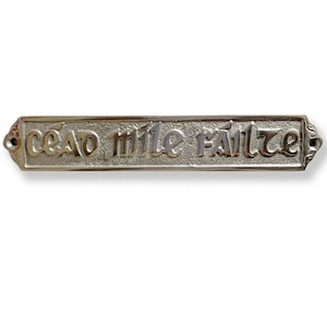 May include: A rectangular, silver-toned metal plaque with the words "cead mile failte" in stylized lettering. The plaque has decorative end pieces and a textured background.