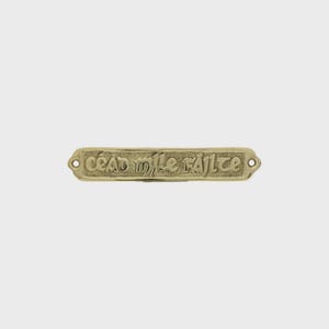 May include: A gold-coloured metal plaque with the embossed text "céad míle fáilte". The rectangular plaque has rounded edges and two holes for mounting. The text is in a stylized font.