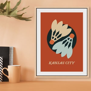 Può includere: Una stampa incorniciata con sfondo rosso che presenta due volanti da badminton stilizzati in un cerchio nero. Il testo "KANSAS CITY" è stampato in bianco sotto i volanti.