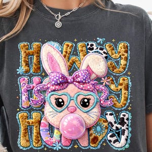 Könnte beinhalten: Dunkelgraues T-Shirt mit einem Cartoon-Hasen, der eine herzförmige Brille trägt und eine Blase bläst. Der Text "Howdy Howdy Howdy" ist in einer verspielten Schriftart mit floralen und Tiermuster-Akzenten gestickt. Der Hase hat eine rosa Schleife.