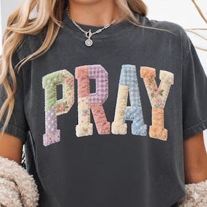 Könnte beinhalten: Dunkelgraues T-Shirt mit dem Wort "PRAY" in bunten Buchstaben im Patchwork-Stil. Jeder Buchstabe hat ein anderes Muster, darunter Blumenstickereien und Vichy-Karo, was ein einzigartiges und strukturiertes Design ergibt. Das Shirt hat einen klassischen Rundhalsausschnitt.