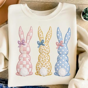 May include: A cream-coloured sweatshirt with three embroidered Easter bunny designs. The bunnies are pink and white checked, yellow leopard print, and blue with white polka dots. Each bunny has a bow.