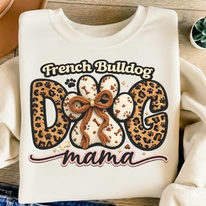 May include: Cream-coloured sweatshirt with the text "French Bulldog Dog mama". The word "DOG" is designed as a paw print with a brown bow. The letters "D" and "G" have a leopard print design.