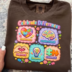 May include: Brown t-shirt with the words "Celebrate Differences" in colorful letters. The shirt features several graphic designs, including a heart-shaped puzzle, a brain, hands forming a heart, a lightbulb, and puzzle pieces. The shirt is a Comfort Colors brand.
