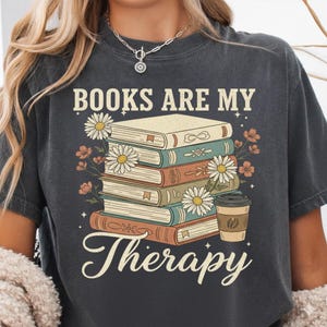 Peut inclure: T-shirt gris foncé avec l'inscription "BOOKS ARE MY Therapy" au-dessus d'une pile de livres, de marguerites et d'une tasse de café. Les livres sont de couleurs variées, et le design a une esthétique vintage.