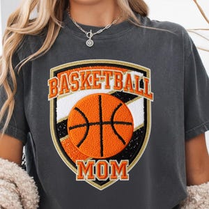 Puede incluir: Camiseta gris oscuro con un gráfico de baloncesto. El gráfico presenta una pelota de baloncesto naranja con líneas negras dentro de un escudo con las palabras "BASKETBALL MOM" en letras naranjas.