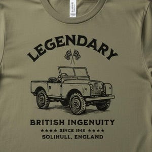 May include: Olive green t-shirt with the words "LEGENDARY" and "BRITISH INGENUITY" printed in black. The shirt features a vintage car illustration and the text "SINCE 1948 SOLIHULL, ENGLAND". The shirt is displayed on a wooden surface.