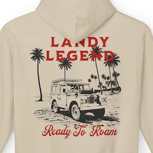 Puede incluir: Sudadera con capucha beige con un gráfico de un vehículo todoterreno vintage, palmeras y el texto "LANDY LEGEND" y "Ready To Roam" en rojo. La sudadera tiene una capucha con cordón y un bolsillo tipo canguro.