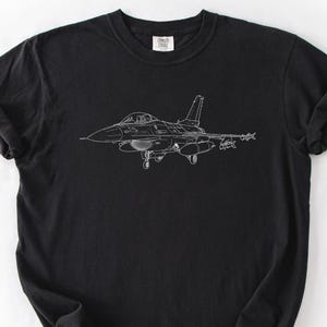 May include: Black t-shirt featuring a white line drawing of a fighter jet. The design shows the jet's silhouette, including the cockpit, wings, and tail. The shirt is made of a soft material and has a classic crew neck.