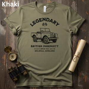 Puede incluir: Camiseta caqui con un gr&aacute;fico de coche antiguo y el texto "LEGENDARY BRITISH INGENUITY SINCE 1948 SOLIHULL, ENGLAND." La camiseta se muestra con una br&uacute;jula, unos prism&aacute;ticos y un pergamino enrollado sobre una superficie de madera.