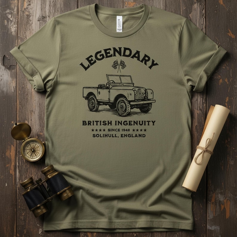 May include: Olive green t-shirt with the words "LEGENDARY" and "BRITISH INGENUITY" printed in black. The shirt features a vintage car illustration and the text "SINCE 1948 SOLIHULL, ENGLAND". The shirt is displayed on a wooden surface.