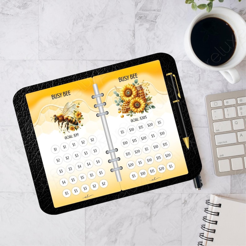 Pu&ograve; includere: Un'agenda in pelle nera con testo "Busy Bee" e illustrazioni di api e girasoli. L'agenda ha pagine per obiettivi finanziari, con importi da 1&euro; a 20&euro;. Sono visibili anche una penna, una tazza di caff&egrave; e una tastiera.