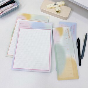 May include: Three pastel colored weekly planner pads with a grid pattern and the text "Weekly Plan" on the top. The pads are on a white surface with two black pens.