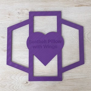 Puede incluir: Corte de corazón morado con el texto "Seatbelt Pillow with Wings" y el texto "Quilted Hugs by LeSha Reitman" en la parte inferior.