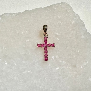 Może przedstawiać: Złoty wisiorek w kształcie krzyża, ozdobiony licznymi małymi, żywymi różowymi kamieniami szlachetnymi. Krzyż jest wyśrodkowany na białym, teksturowanym tle, podkreślając wzór i kolor wisiorka.