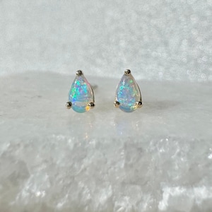 Puede incluir: Un par de pendientes de ópalo con forma de lágrima. Los ópalos muestran un vibrante juego de colores, engastados en metal dorado. Los pendientes se exhiben sobre una superficie blanca texturizada.