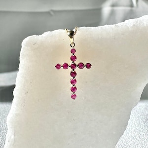 Pode incluir: Um pingente de cruz de ouro com pedras de rubi vermelho. A cruz está sobre um fundo branco.