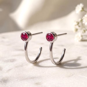 May include: A pair of silver-toned hoop earrings featuring a small, round, faceted red gemstone set in a bezel at the top of each hoop. The earrings have a post-back closure and a polished metallic finish, reflecting light on their curved surfaces.