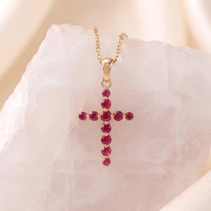 Puede incluir: Un collar de oro con un colgante de cruz engastado con pequeñas piedras redondas de color rojo intenso. La cruz está compuesta por once piedras individuales dispuestas en un patrón vertical y horizontal, suspendida de una delicada cadena de oro.