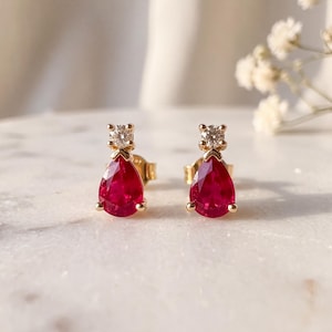 May include: A pair of gold stud earrings, each featuring a pear-shaped red gemstone suspended beneath a small, round, clear faceted stone. The earrings are crafted with a polished gold setting, showcasing a vibrant red hue and a brilliant sparkle.