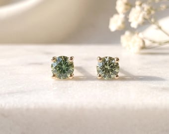May include: A pair of stud earrings featuring round-cut, olive-green gemstones set in four-prong gold metal settings. The earrings are positioned side-by-side, showcasing the faceted surface of the stones and the metallic structure of the prongs.