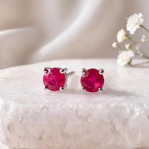 May include: A pair of stud earrings featuring round, faceted, vibrant red rubies set in four-prong silver-toned metal settings. The earrings are positioned side by side, showcasing the deep crimson color and the reflective quality of the stones.