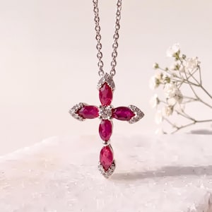 May include: A silver cross pendant necklace featuring four marquise-cut deep pink rubies arranged in a cross shape, accented with small clear round-cut stones at the center and along the edges of the metal frame, suspended on a delicate silver chain.