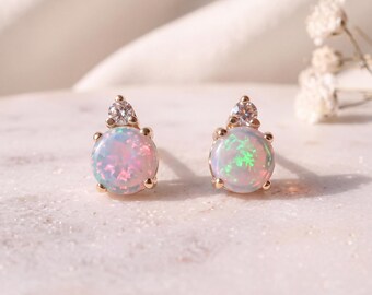 Op de afbeelding: Een paar oorknopjes met een ronde, iriserende opaal cabochon gezet in goud, bekroond met een kleine, heldere, rond geslepen edelsteen. De opalen vertonen flitsen van roze, groen en blauw licht, vastgezet met vier pootjes in een gouden zetting.