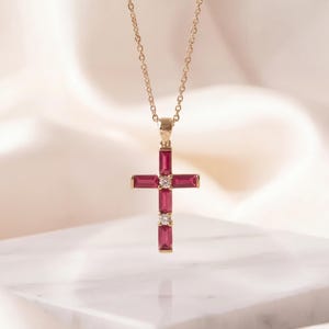 May include: A gold necklace featuring a cross pendant set with baguette-cut red gemstones and two small round clear stones at the center and lower section. The pendant hangs from a delicate gold chain, showcasing a refined and structured design.