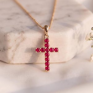 Può includere: Una collana color oro con un ciondolo a forma di croce incastonato con molteplici piccole pietre rotonde di un rosso vibrante. Il ciondolo pende da una delicata catenina color oro, mostrando un design classico con finitura metallica lucida e accenti rossi.