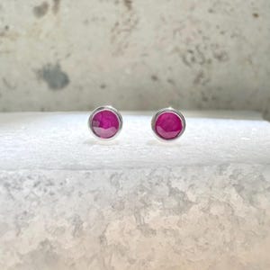 May include: A pair of stud earrings featuring round, deep pink gemstones set in silver-colored metal. The earrings are displayed on a textured, white surface, showcasing their polished appearance. The gemstones have a faceted cut.