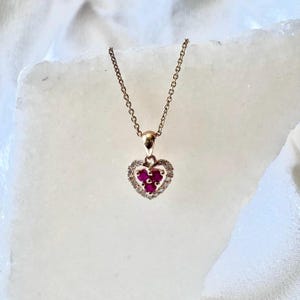 May include: A rose gold necklace with a heart-shaped pendant. The pendant features a cluster of red gemstones surrounded by a halo of small, clear stones. The necklace is displayed on a white surface.