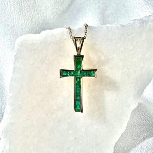 May include: A gold cross pendant featuring a series of square-cut green emeralds set in a vertical and horizontal arrangement, suspended from a delicate gold chain. The cross has a polished gold frame that surrounds the vibrant green gemstones.