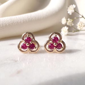 May include: A pair of gold stud earrings, each featuring three round, deep pink rubies arranged in a cluster. The stones are set within a polished gold frame that forms a three-lobed, clover-like shape, creating a cohesive and symmetrical design.