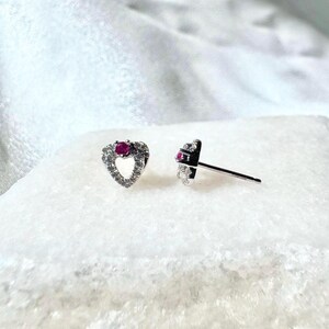 May include: A pair of heart-shaped stud earrings. Each earring features a small, round, red gemstone at the center of the heart. The heart is outlined with small, clear gemstones. The earrings are set on silver posts.