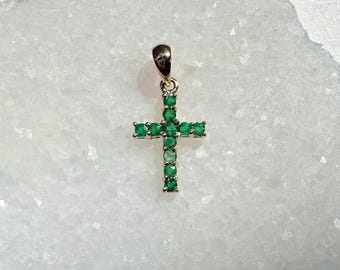 May include: A gold-colored pendant in the shape of a cross, adorned with numerous small, vibrant green gemstones. The cross is centered in the frame, set against a white, textured background. The pendant's design is simple yet elegant.