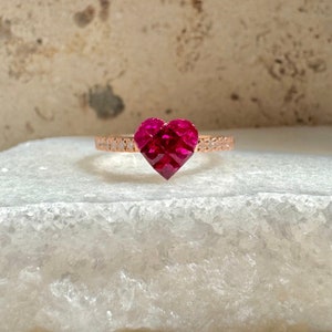 May include: A rose gold ring featuring a faceted, heart-shaped, deep red gemstone. The band is adorned with small, clear gemstones. The ring is set against a neutral background, highlighting the vibrant color of the heart-shaped stone.